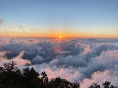 A stunning sunrise over a sea of clouds, with the orange sun breaking through the horizon, casting a warm glow on the landscape below.