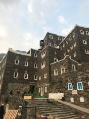 A towering, traditional stone building with many windows and a unique architectural style stands beneath a cloudy sky in the Asir region.