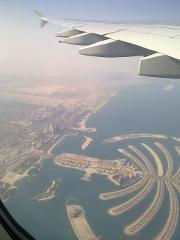Aerial view of Dubai's Palm Jumeirah and skyline from an airplane window, with the plane's wing visible in the upper right.