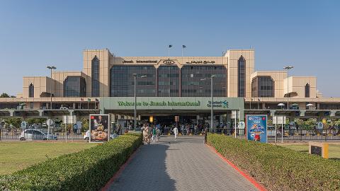 This is a daytime shot of the Jinnah International Airport terminal in Karachi, Pakistan, with signage in English and Arabic, showing the entrance with people and advertisements.
