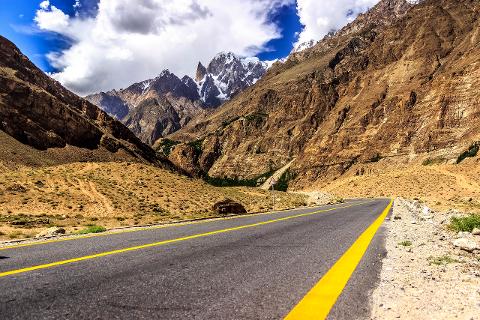 A road winds through a mountainous landscape under a blue sky with fluffy clouds, showcasing a stunning natural environment.
