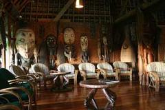 A warmly lit room at Karawari Lodge features carved wooden statues and masks adorning the walls, complemented by comfortable chairs and small wooden tables on a polished wooden floor.