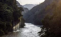 A lush river valley with a waterfall on the left side and mountains in the background, some birds flying in the sky.