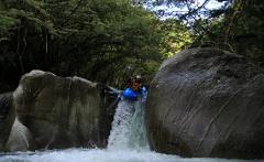 A kayaker in a blue kayak paddles down a small waterfall between two large boulders in a lush, green forest.