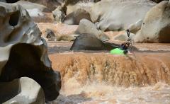 A kayaker navigates a rushing, muddy river filled with large rocks, using a paddle to steer through the turbulent water.