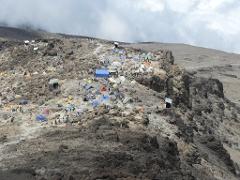 A crowded campsite scene on Mount Kilimanjaro, with tents scattered across rocky terrain under a cloudy sky.