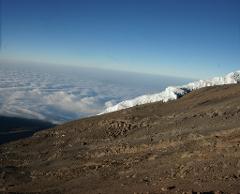 A high-angle view shows a rocky, brown mountainside leading to a snowy peak, with a sea of clouds visible in the distance under a blue sky.