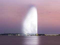 A tall fountain of water shooting up into the air in front of a city skyline during dusk with a pastel sky.