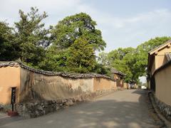 A road winds between traditional Japanese buildings with textured walls, topped with trees under a bright sky.