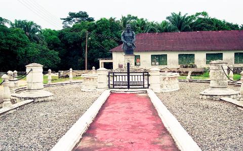 A statue of a man stands in front of a building with a red roof, surrounded by trees and a decorative fence with a red walkway leading to it.