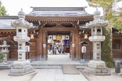 A serene view of a traditional Japanese shrine entrance with stone lanterns and a wooden gate, leading to a courtyard with visitors.
