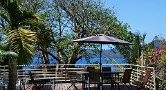 A patio with a table and chairs under an umbrella overlooks a stunning view of the ocean and a lush green landscape.