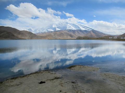 A serene mountain landscape with a calm lake reflecting the sky and clouds, bordered by barren hills and snow-capped peaks.