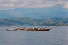 A panoramic view of fishing boats with fishermen on a tranquil lake against a backdrop of rolling green hills and a cloudy sky.