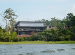 The image shows the Lake Murray Lodge, a stilted building on a lush, green shoreline with trees, viewed from the water, possibly a lake or river.