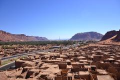 A wide-angle shot reveals a sprawling desert landscape featuring an ancient mud-brick town, a modern road, palm groves, distant mountains under a clear blue sky.