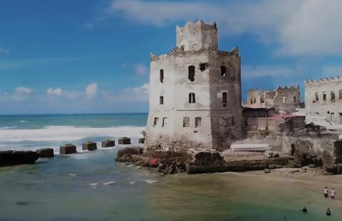 Tur privat de 3 zile în Mogadiscio (istorie, cultură și coastă) An old, weathered, multi-story building stands on a beach, with waves crashing against the rocks and people on the sand.