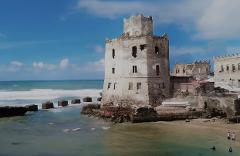 Tur privat de 3 zile în Mogadiscio (istorie, cultură și coastă) An old, weathered, multi-story building stands on a beach, with waves crashing against the rocks and people on the sand.