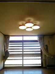 A sunlit room with a patterned ceiling features a four-light ceiling fixture and large horizontal blinds covering a window, with curtains draped on either side.