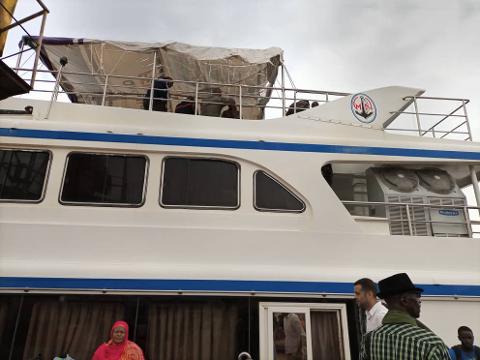 A white boat with blue stripes is shown, with people standing around it, some on the top deck under a makeshift awning.