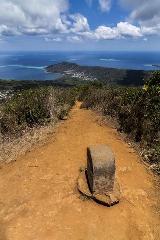 A dirt trail leads from a stone marker towards a scenic overlook with blue water, lush green hills, and a cloudy sky.