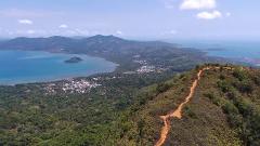 A scenic view from a mountain ridge with a winding path, overlooking a coastal town, lush forests, and calm blue waters.