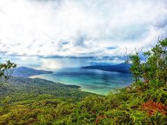 A scenic view of a lush, green island landscape overlooking a turquoise sea under a cloudy sky, captured from an elevated vantage point.