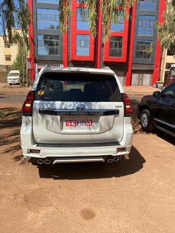 A white Land Cruiser Prado is parked on a dirt lot in front of a red and gray building with many windows.
