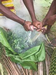 The image shows people preparing food, likely fish and vegetables, wrapped in banana leaves with steam rising from the cooking process.