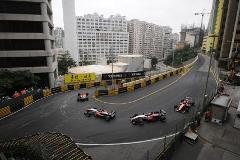 A high-angle shot captures Formula 3 cars racing on the tight Guia Circuit in Macau, lined with barriers and surrounded by urban buildings.