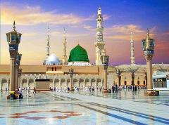 A grand mosque with a green dome and minarets stands prominently in a vast, sunlit plaza filled with people.