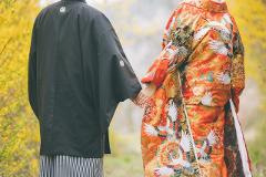 A couple in traditional Japanese wedding attire walks hand-in-hand, with the groom in a black kimono and the bride in a vibrant orange and white crane-patterned kimono.