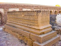 An intricately carved stone tomb stands in front of brick arches and a wall, likely a historical or religious site.