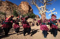 Dogon people performing a traditional dance in Mali, wearing colorful masks and costumes. The backdrop features unique rock formations and a clear blue sky.