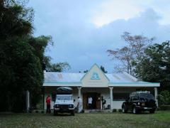 A white building with "CENTRE D'INTERPRETATION" written on it stands surrounded by trees, with two parked vehicles and several people standing in front of it.