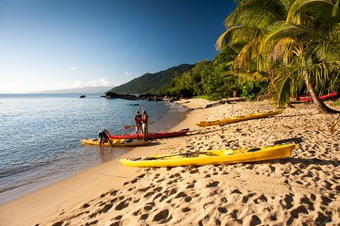 A sunny beach scene with kayaks on the sand, people preparing to kayak, lush green trees and a mountain range in the background.