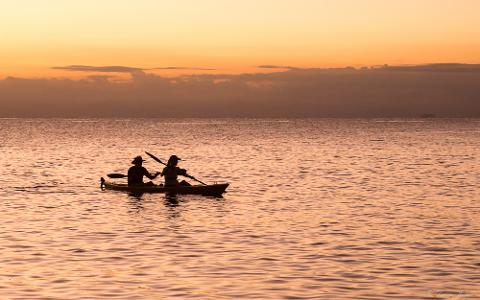 Silhouette of two people paddling in a kayak on calm water at sunset with a hazy sky.