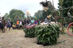 Two large, leaf-covered figures with cone-shaped, decorated heads stand in a grassy area, surrounded by onlookers.