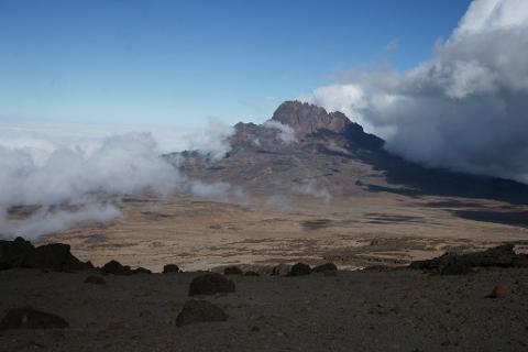 A breathtaking view of Mount Kilimanjaro's rugged peaks peeking through swirling clouds, set against a vast, arid landscape under a blue sky.