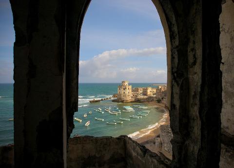 Tur privat de 3 zile în Mogadiscio (istorie, cultură și coastă) An arched window frame reveals a coastal scene with a building, beach, turquoise water dotted with boats, and a blue sky with fluffy clouds.