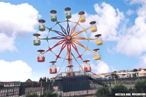 Tur privat de 3 zile în Mogadiscio (istorie, cultură și coastă) A colorful Ferris wheel stands tall against a blue sky with scattered clouds, atop a structure behind a green fence, adding a whimsical touch to the landscape.