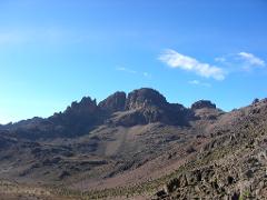 A mountainous landscape dominates the frame, showcasing rugged peaks under a clear blue sky with scattered clouds.