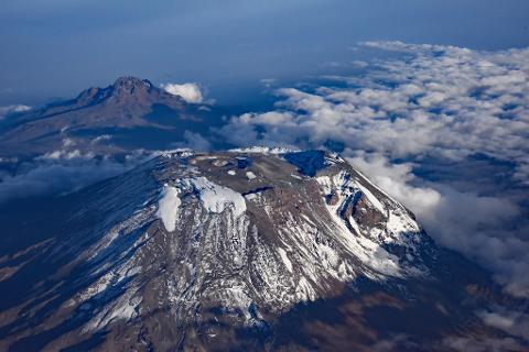 An aerial view shows snow-capped Mount Kilimanjaro and Mount Meru rising above a sea of clouds under a blue sky.