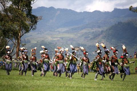 A line of tribesmen in traditional attire, adorned with headdresses, body paint, and skirts, process across a grassy field with mountainous scenery in the background.