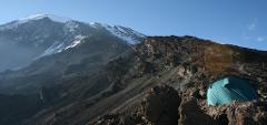 A tent sits nestled amidst rocky terrain on a mountain slope, with a snow-capped peak looming in the background under a clear blue sky.