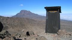 A rugged mountain landscape featuring a wooden outhouse labeled "Porters Toilet" on a rocky outcrop, set against a backdrop of distant peaks and a sea of clouds under a clear blue sky.