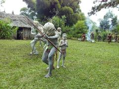 A group of people dressed as warriors in traditional costumes and masks, stand on a grassy field near a hut.
