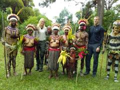 A group of people, including tourists and locals dressed in traditional attire with spears and headdresses, pose for a photo outdoors on a grassy field.