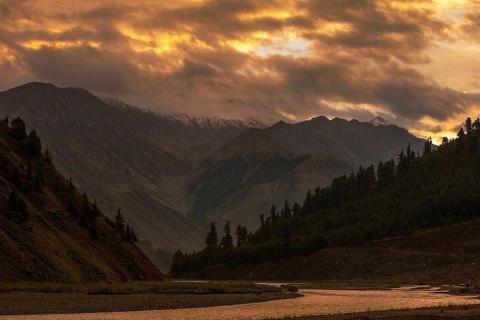 A serene mountain landscape featuring a golden sunset with clouds, a river flowing through a valley, and forested slopes.