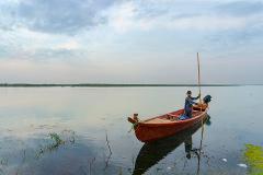 A young boy stands in a red boat on a calm lake, holding a long pole under a cloudy sky.